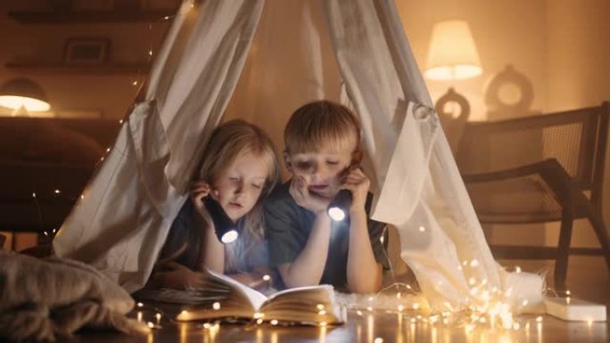 小孩子在客厅的毯子上用手工帐篷用手电筒看书