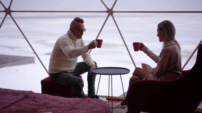 侧视图自信的男人和女人在冬天坐在玻璃露台上喝咖啡敬酒。轻松快乐的高加索成年夫妇享受周末休闲。