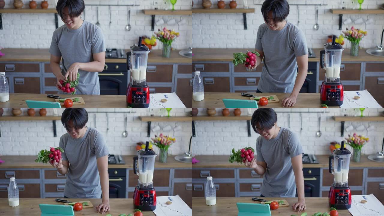 开朗的亚洲男人，露齿微笑，烹饪健康的沙拉，站在厨房台面的平板电脑摄像头上聊天。积极微笑的千禧一代准备