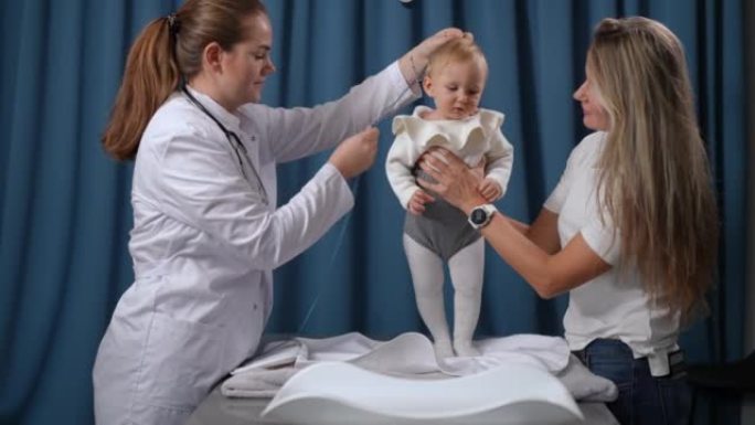 漂亮的女婴站在桌子上的肖像，母亲抱着孩子和医生测量身高。无忧无虑可爱的快乐高加索蹒跚学步的孩子在室内