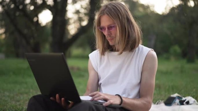 中等镜头集中的白人年轻人在笔记本电脑上上网，想着放下太阳镜看着设备屏幕。自信英俊的千禧一代在春夏公园