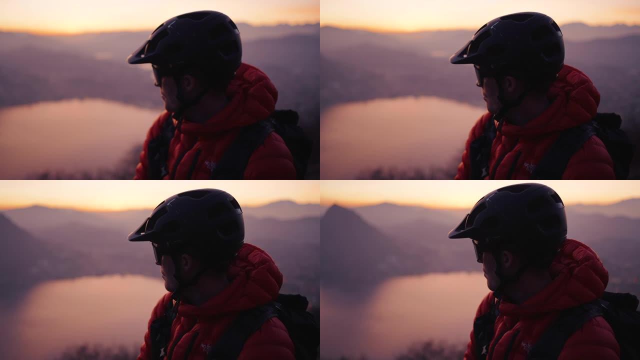 山地车手坐在山顶上，在日落时眺望下方的城市