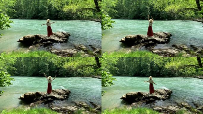 年轻漂亮的女人穿着长裙和草船帽站在石头上，靠近强大的涟漪山河，跳舞。夏季旅游概念，浪漫复古女孩，旧复
