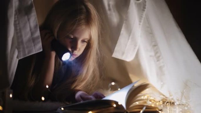 一个可爱的小女孩晚上在黑暗中坐在带花环的帐篷里的房子里用手电筒读一本童话故事。