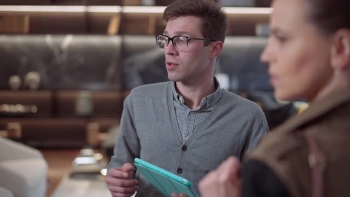 自信的年轻男性卖家在眼镜上的肖像，平板电脑站在家具店里与客户交谈。专家高加索推销员在室内豪华商店陈列