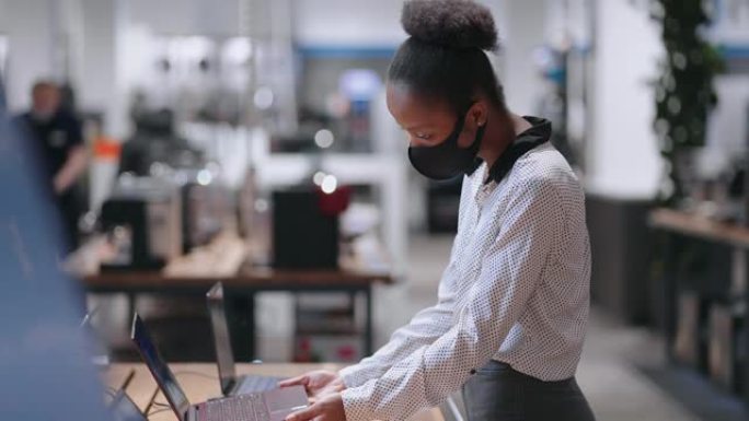 戴着口罩的年轻非洲裔美国女性正在电子商店选择现代笔记本电脑，观看展览样本