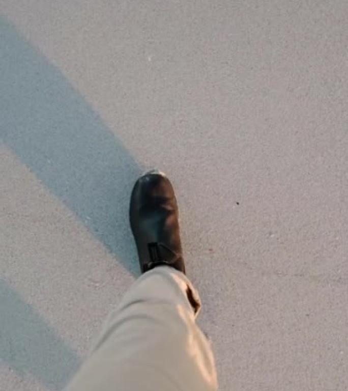男人的腿穿着裤子和鞋子在沙滩上行走的第一人称视角