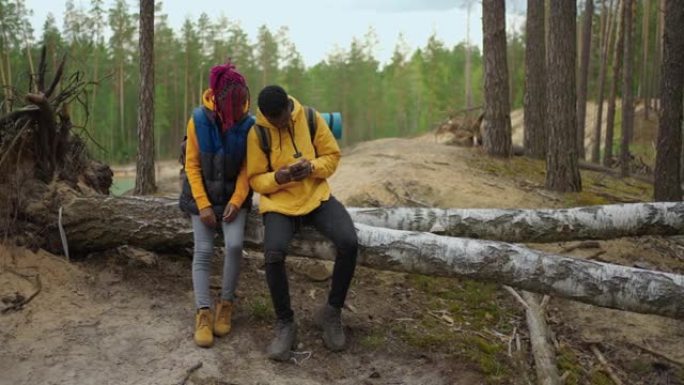 一个黑人和一个女人坐在原木上，看着山林中的自然之美。花时间在一起，用你的智能手机导航。绘制路线