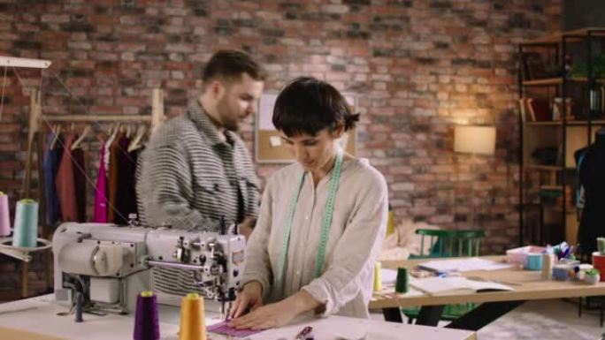 一个短发的女人在缝纫机上工作，而她身后的一个男人在桌子上工作，接听电话并写下设计师的笔记。在阿里·阿