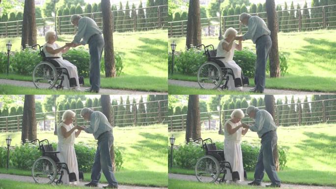 在阳光明媚的夏日公园，高级男子帮助美女从轮椅上站起来。慈爱的丈夫支持白人妻子。事故和康复概念。