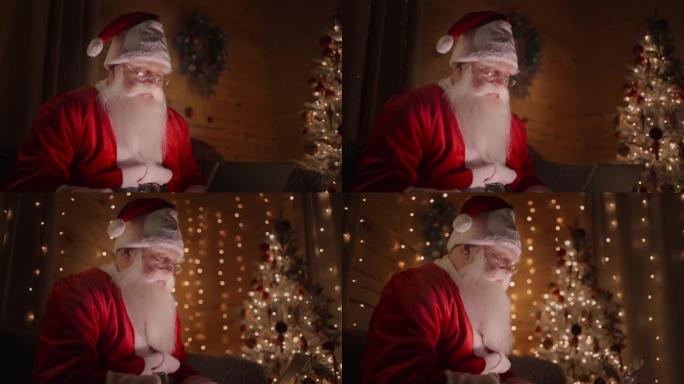 圣诞老人正在家里的电脑上工作，戴着眼镜，在装饰好的圣诞树的背景上留着胡须，神奇的圣诞灯