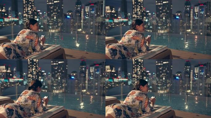 傍晚时分，一名亚裔女子拿着手机坐在高楼的游泳池里，俯瞰着这座城市。