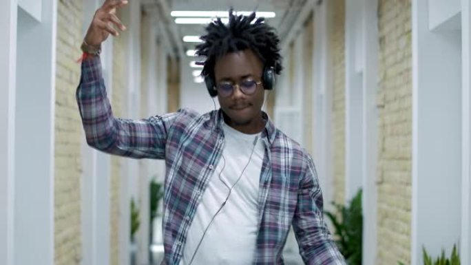穿着眼镜和耳机的轻松非洲裔美国年轻人在办公室走廊跳舞。快乐自信的员工在工作场所玩得开心的肖像。电影院
