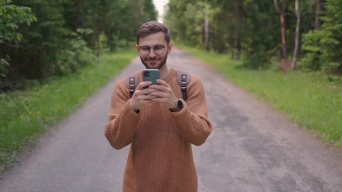 一个背着背包的男人沿着树林中的一条土路走，写信息，在线交流，查看内容，微笑，欢欣鼓舞。前视图。