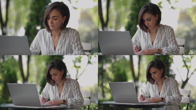 学生女子布鲁内特阿拉伯西班牙裔民族通过互联网远程学习，坐在夏季咖啡馆里拿着笔记本电脑，写下笔和笔记本