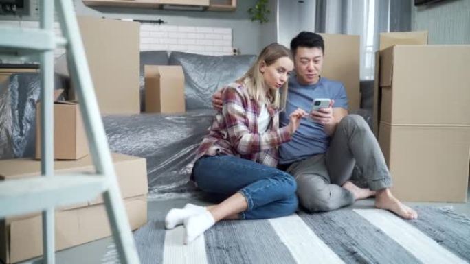 年轻的家庭在一个新的公寓里，纸板箱坐在地板上，愉快地选择新家具网上商店，使用智能手机应用程序购物。
