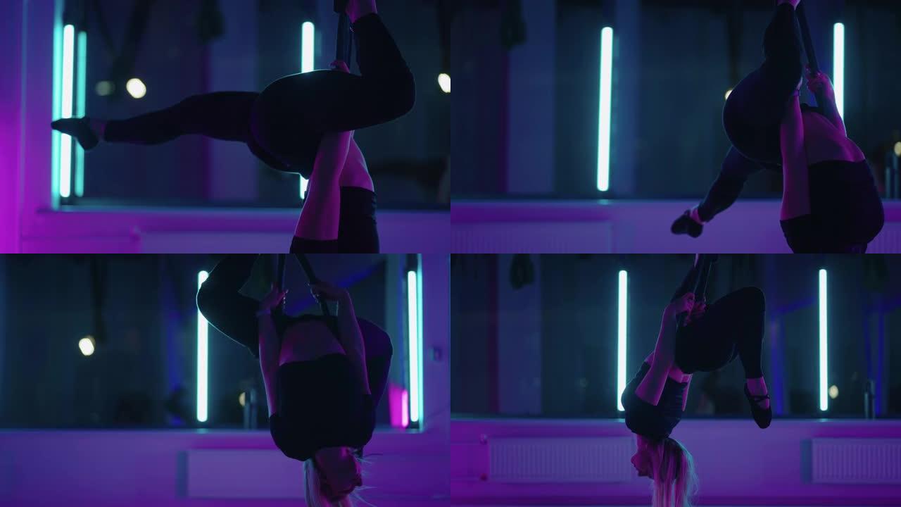 一个女人在霓虹灯下做瑜伽并在悬吊的吊床上伸展。一个女人在工作室的吊床上飞，做伸展运动和身体锻炼。空中