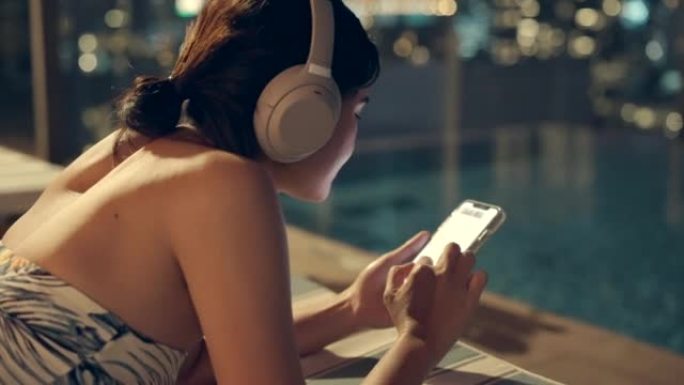 她听音乐，用手机玩网上购物。泰国曼谷城景高楼上的游泳池区。