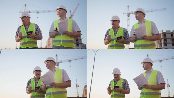 两名穿着安全服的中年男性建筑工人的腰部站立在施工现场，男子使用对讲机，他的同事拿着带有项目计划的纸