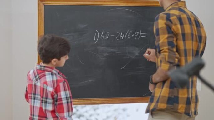 中东老师在黑板上写算术表达式，并将粉笔传给解决数学问题的小男孩。集中男孩在教室学习数学的后视图。