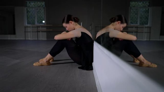 侧视图沮丧的疲惫的芭蕾舞女演员坐在镜子的地板上，将头放在膝盖上。疲惫沮丧的年轻白人妇女在舞蹈室叹息。