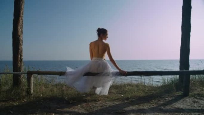 苗条的芭蕾舞演员在海河黎明时晴朗的夏日天空背景下的后视图。优雅优雅的高加索女人，穿着芭蕾舞短裙，坐在