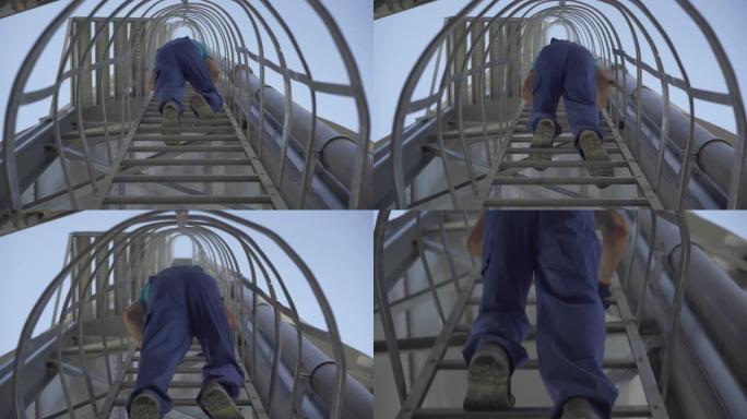 穿着蓝色制服的工厂工人在巨大的存储塔上爬下梯子的仰视图。无法辨认的高加索人在户外的水泥筒仓上，背景是