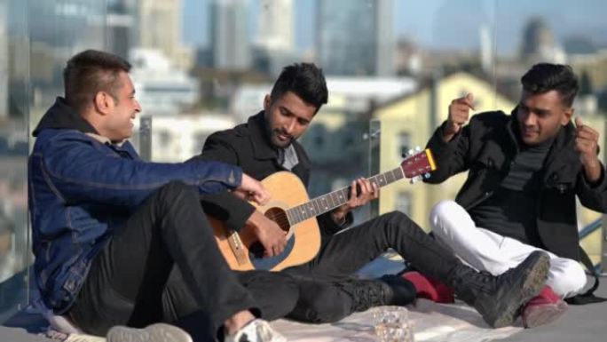 宽幅自信放松的男人弹吉他，朋友们坐在阳光下的屋顶上跳舞。快乐无忧无虑的中东男人在城市阳光明媚的日子享