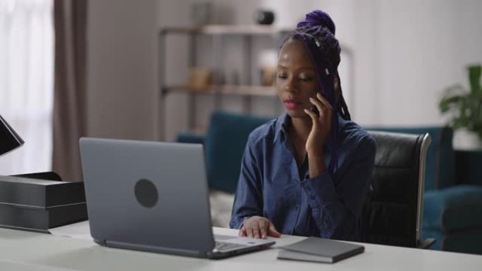 女销售经理在家庭办公室工作，黑人妇女通过手机打电话，坐在笔记本电脑旁，咨询客户