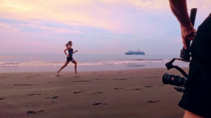 摄影操作员拍摄赤脚在海滩上奔跑的女人。慢动作，日落景观