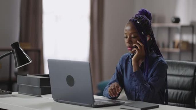 黑人妇女正在用手机打电话，坐在家里办公室的工作台上，用手机聊天，中等肖像