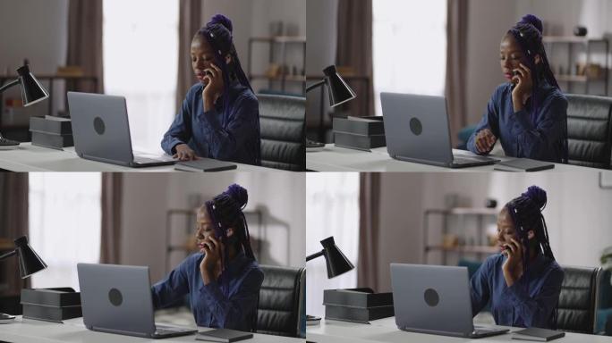 黑人妇女正在用手机打电话，坐在家里办公室的工作台上，用手机聊天，中等肖像