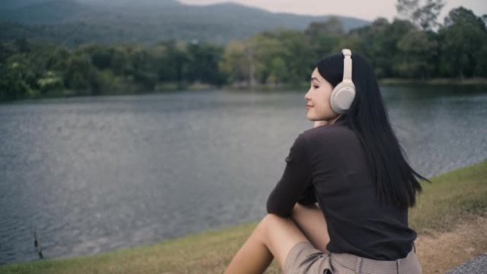 亚洲女孩戴着耳机听音乐，脸上挂着快乐的微笑，坐在公园的座位上，有湖泊和山脉。