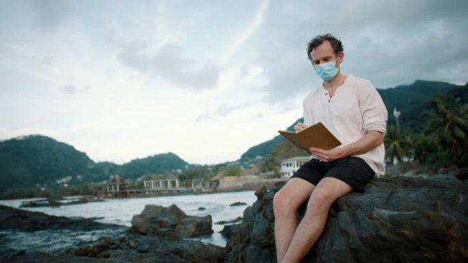 一名戴着面具的男子坐在海洋附近的岩石上写书的特写镜头