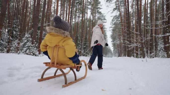 快乐的年轻女子和她的小孩在冬天的森林里散步，儿子坐在木制雪橇上