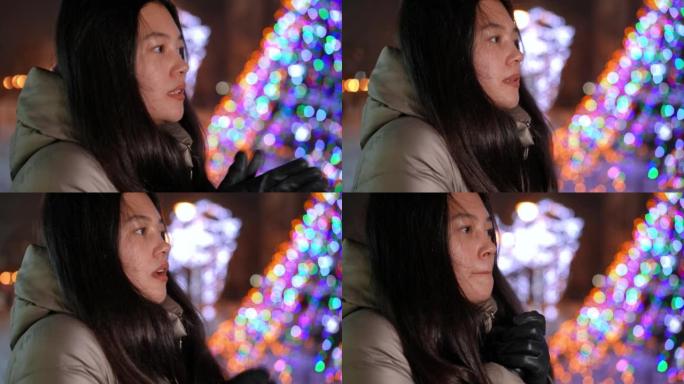 侧视特写冻结的亚洲女孩站在户外圣诞晚会上吹手。青少年在除夕夜在城市城市街道上等待，背景照明。