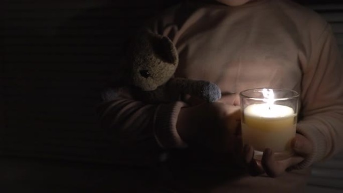 窗边黑暗房间里的一个女孩手里拿着蜡烛和泰迪熊