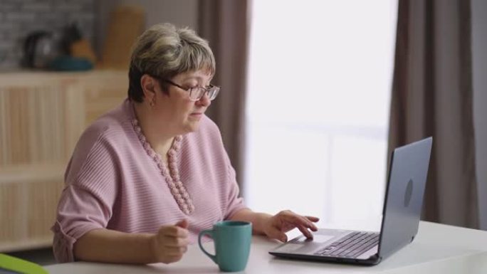 祖母在厨房使用笔记本电脑，戴着眼镜的老年妇女正在喝茶和上网