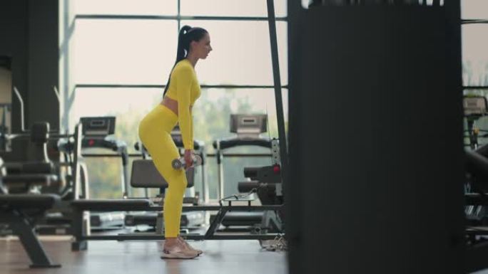 适合西班牙裔女性穿着黄色运动服在健身房用哑铃进行举重硬拉运动。女人黑发健身表演用哑铃做硬拉运动
