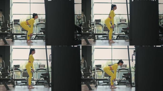 适合西班牙裔女性穿着黄色运动服在健身房用哑铃进行举重硬拉运动。女人黑发健身表演用哑铃做硬拉运动