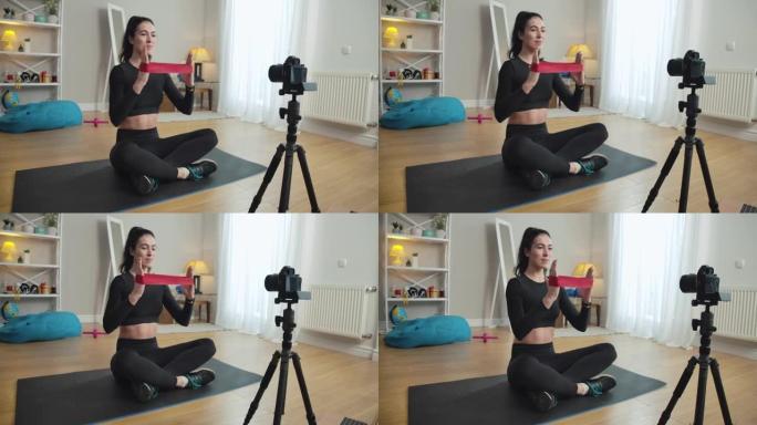 微笑的女运动员用阻力乐队训练双手，在家录制视频博客。苗条自信的白人妇女在室内锻炼和写博客的肖像。电影