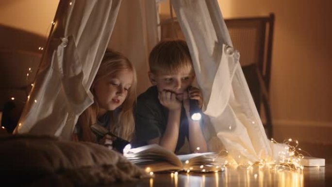 金发小女孩和她的哥哥在帐篷里的黑暗房间里看书，手电筒闪闪发光