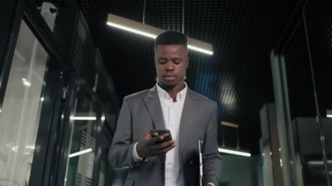 一名黑人商人拿着手机走在走廊上，看着屏幕