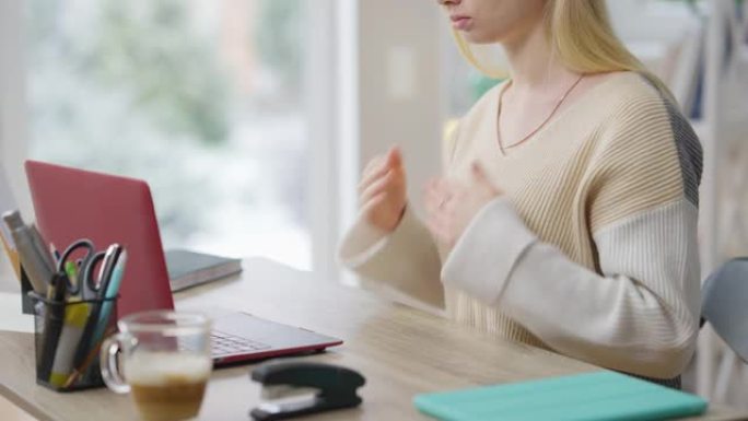 无法辨认的苗条金发女人用手语打手势坐在笔记本电脑的桌子上。严重集中的高加索聋哑女士在网上学习或视频聊