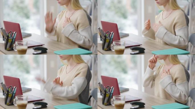 无法辨认的苗条金发女人用手语打手势坐在笔记本电脑的桌子上。严重集中的高加索聋哑女士在网上学习或视频聊