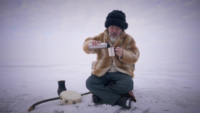 大胡子的蒙古人坐在户外白雪皑皑的冰上慢动作在杯子里倒茶的宽镜头肖像。免费土著老人享受休闲，背景为冬季