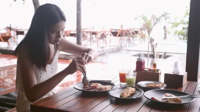 亚洲女孩在海边吃早餐，在平底锅上吃美式早餐，早上吃鸡蛋煎蛋卷培根香肠新鲜食物。