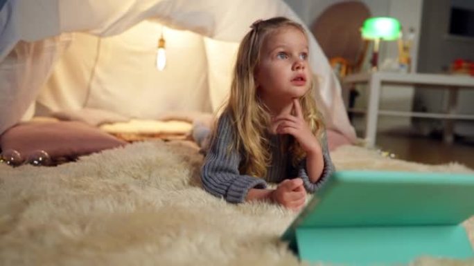 迷人美丽的小女孩在客厅的帐篷里仰望平板电脑。古玩无忧无虑快乐的高加索孩子在室内休息看动画片的宽镜头肖
