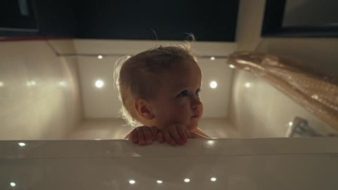 金发碧眼的蹒跚学步的孩子坐在浴缸里，顶着天花板上的明亮灯光