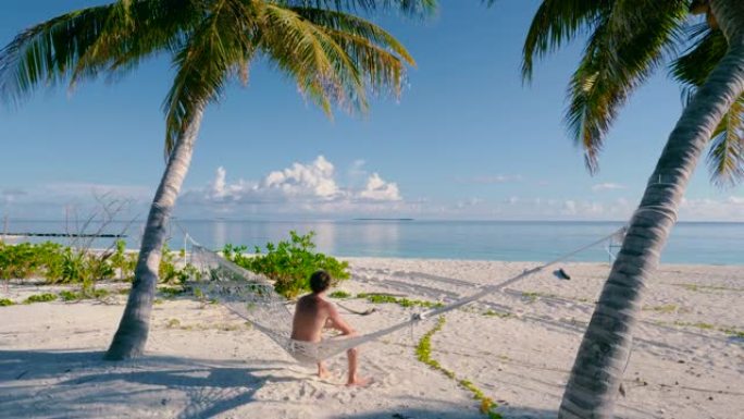 男人坐在海滩上两棵棕榈树之间的吊床上